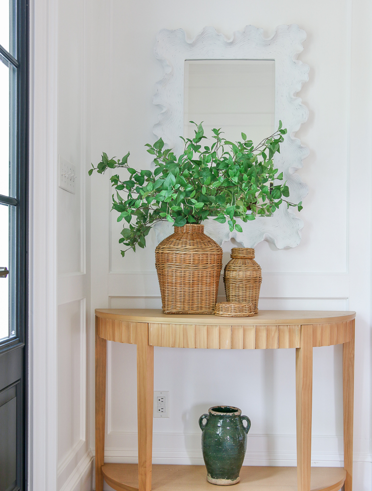 entry with white walls and black door, rounded and fluted small console table with vase on bottom shelf, set of wicker jars on surface with faux greenery styled  white mirror above console