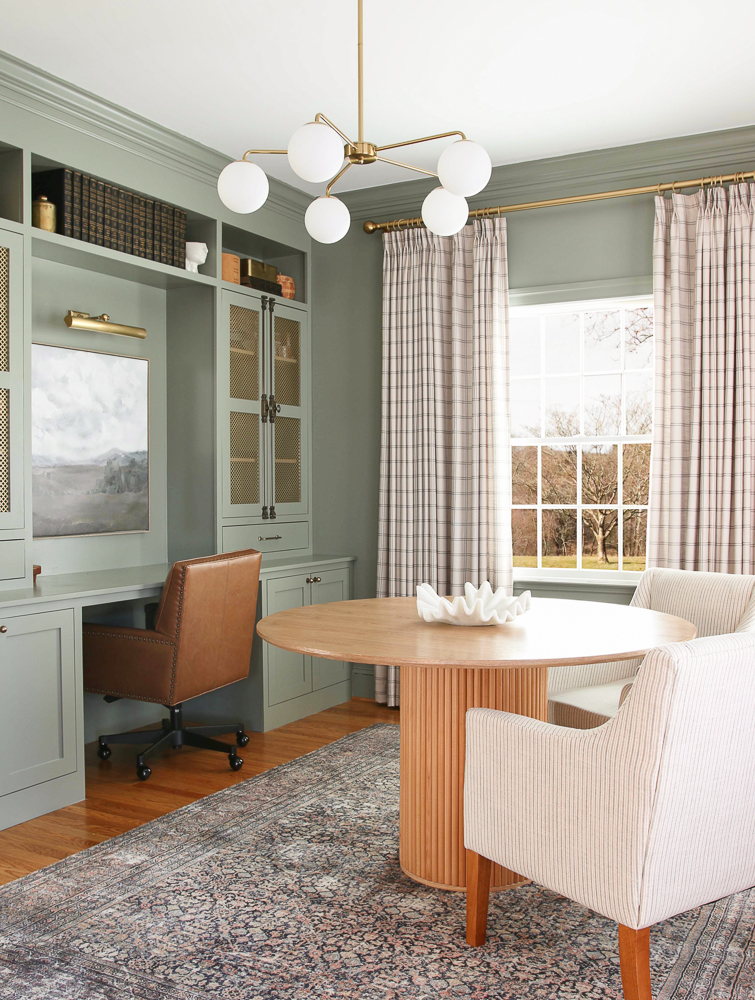 home office painted Storm Cloud Gray, builtin cabinets and desk, brass drape rod and chandelier, Stefana Silber x Two Pages drapes, antique brass hardware, round table centered in room with two upholstered chairs, leather desk chair, grayish green walls