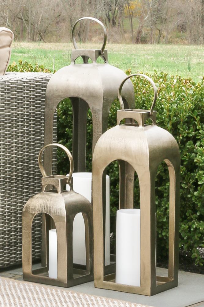 three varying sized gold outdoor metal lanterns with battery operated candles on a bluestone patio next to an outdoor sofa