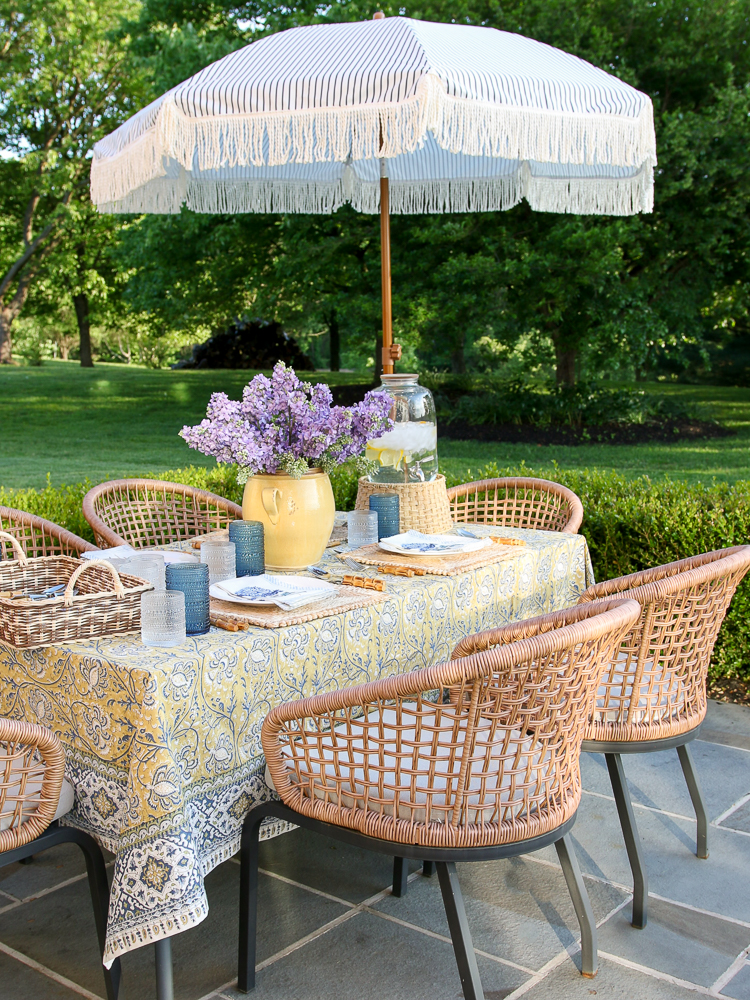 dining area of outdoor entertaining area, rectangular table with yellow printed pottery barn table cloth, woven chairs, umbrella off to side, blue and white tablescape, bluestone patio
