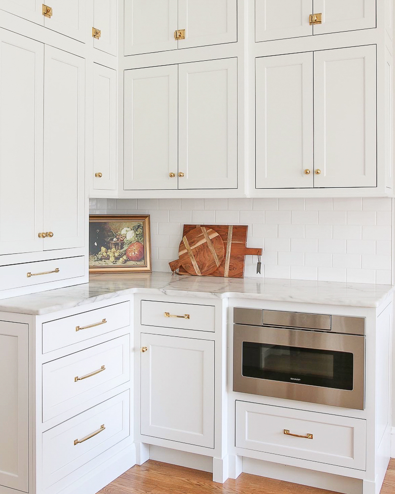 kitchen with factory painted white cabinets, honed marble countertops, unlacquered brass hardware, wood floors, artwork and cutting boards on countertop, drawer microwave