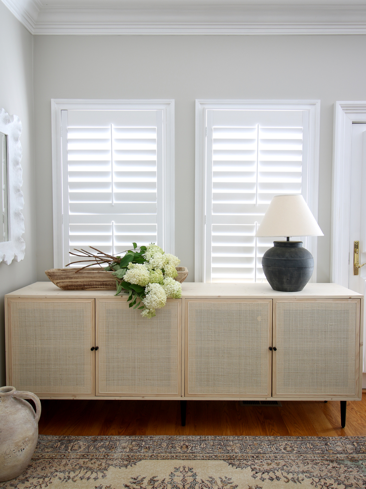 cane cabinet with clear natural stain in front of windows with shutters and a oriental rug. Black ceramic lamp.