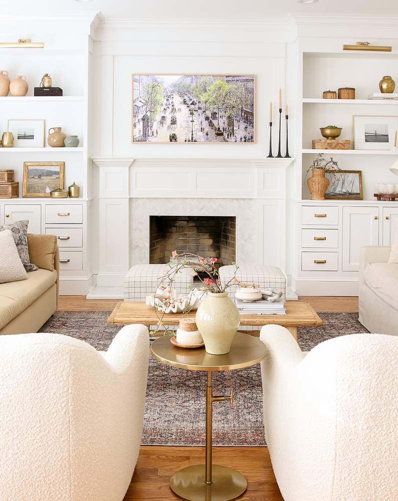 bright white living room with frame tv above fireplace and built-in shelves on fireplace wall, boucle chairs, vintage coffee table, velvet sofas, Loloi rug, brass hardware, picture lights