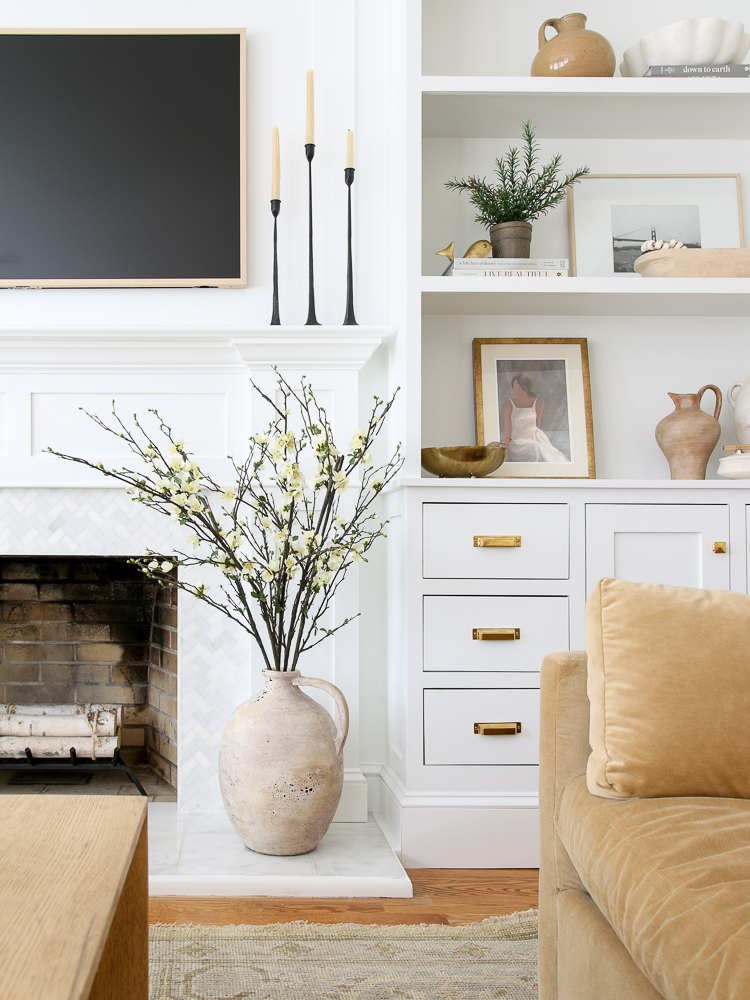 white fireplace wall with frame tv above and builtin shelves and cabinets to the right, shelf decor, vase with branches on floor to the right of the fireplace, velvet sofa