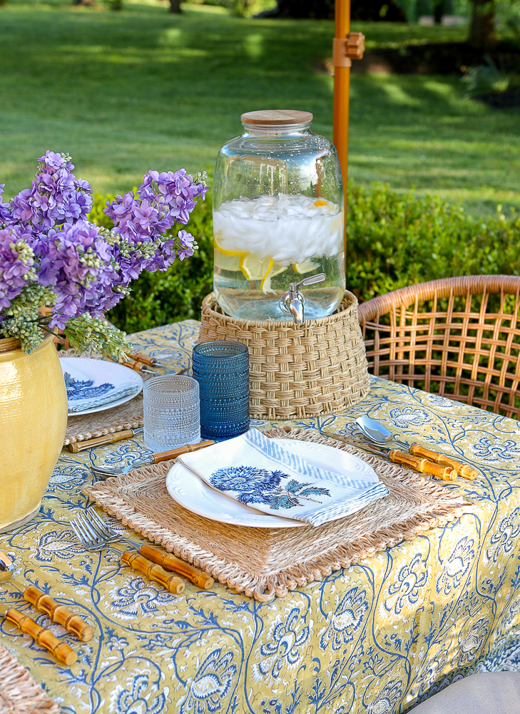 outdoor entertaining area with block print tablecloth, woven chargers, white dishes, blue and clear hobnail glasses, drink dispenser on a woven stand, yellow vase with lilacs, 