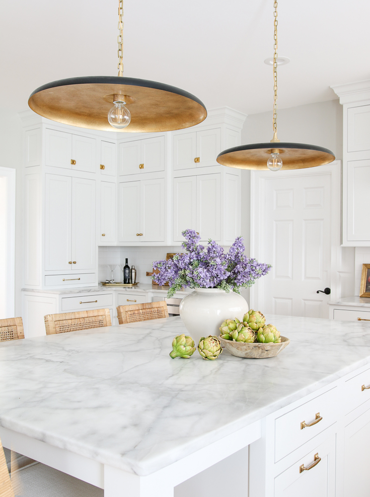kitchen trend of white cabinets, honed marble countertops, pendants over island, rattan counter height stools, vase with faux lilacs, wooden bowl with faux artichokes