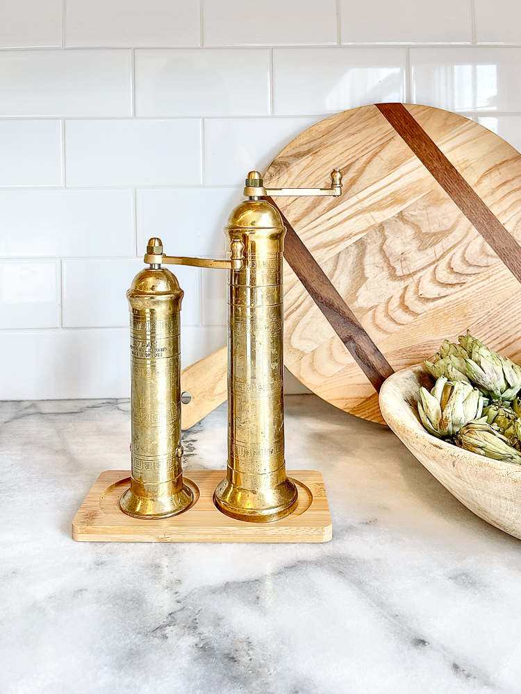 timeless kitchen trend with white staggered subway tile, honed marble countertops, brass mills, round cutting board, wooden bowl with faux artichokes