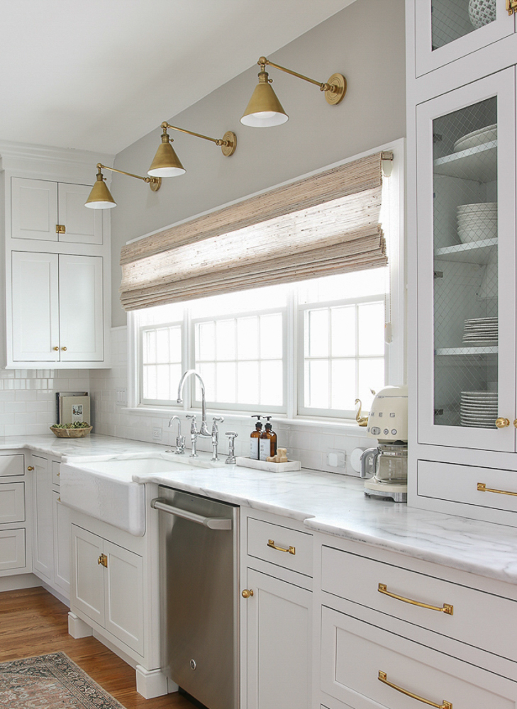classic white kitchen with triple window over the sink, Willowbloom Home woven shade, honed marble countertops, unblacquered brass cabinet hardware, hardwood floors, white subway tile, SW Agreeable Gray