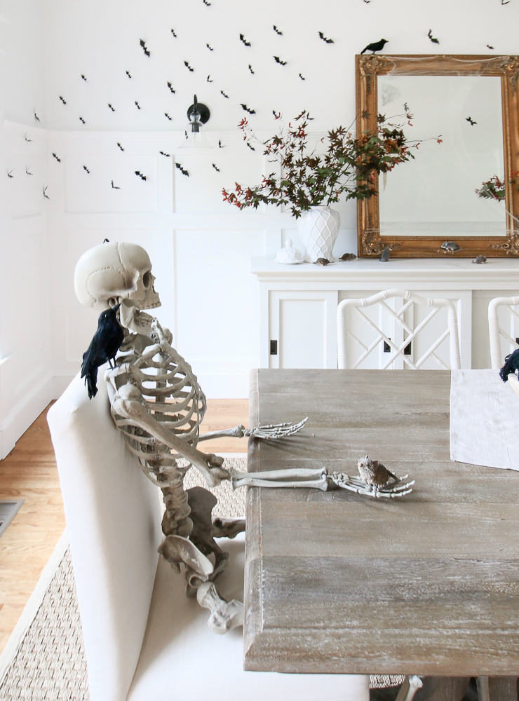 white dining room decorated with classy halloween decor, ideas include skeleton sitting at table with blackbird perched on shoulder, bat colony on wall