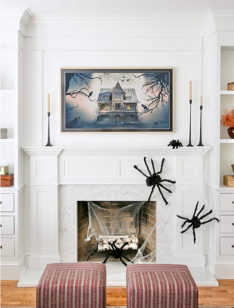 living room fireplace wall with oversized scary spiders, frame TV with haunter house art, candles, ottomans, classy halloween decorating idea for living room fireplace wall. 