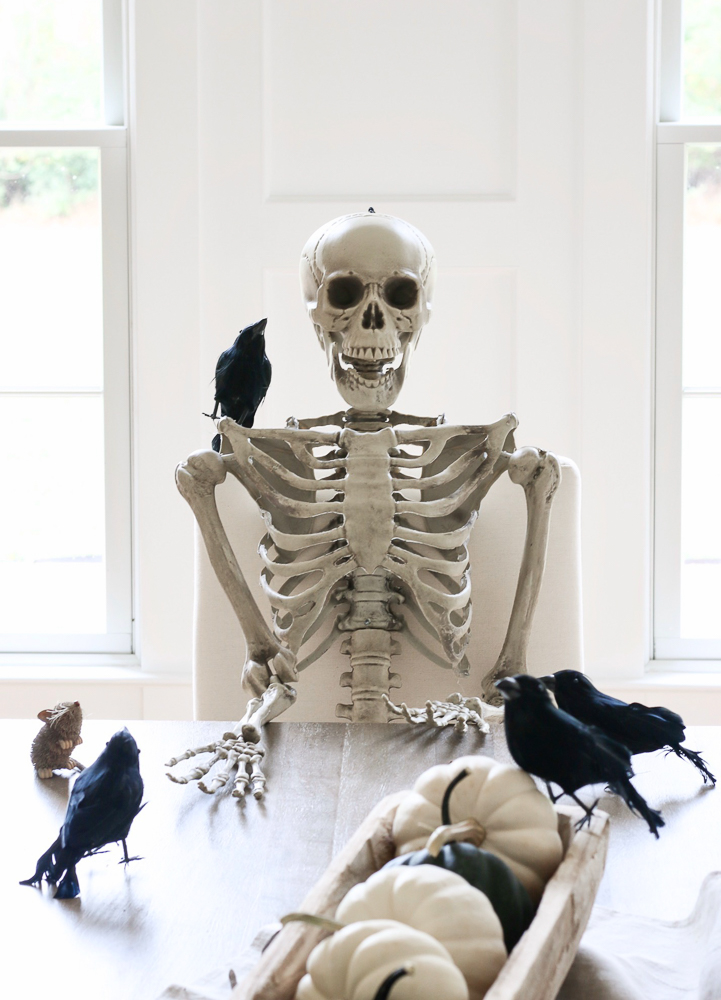 classy halloween decorating ideas showing skeleton sitting at dining table with blackbird perched on shoulder