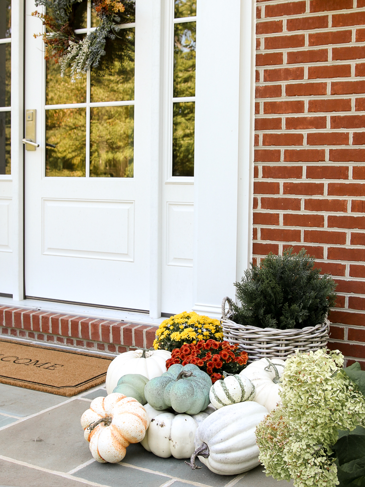 close up of fall porch decor, faux pumpkins, moms, and faux greenery in a rattan planter, welcome mat on bluestone porch