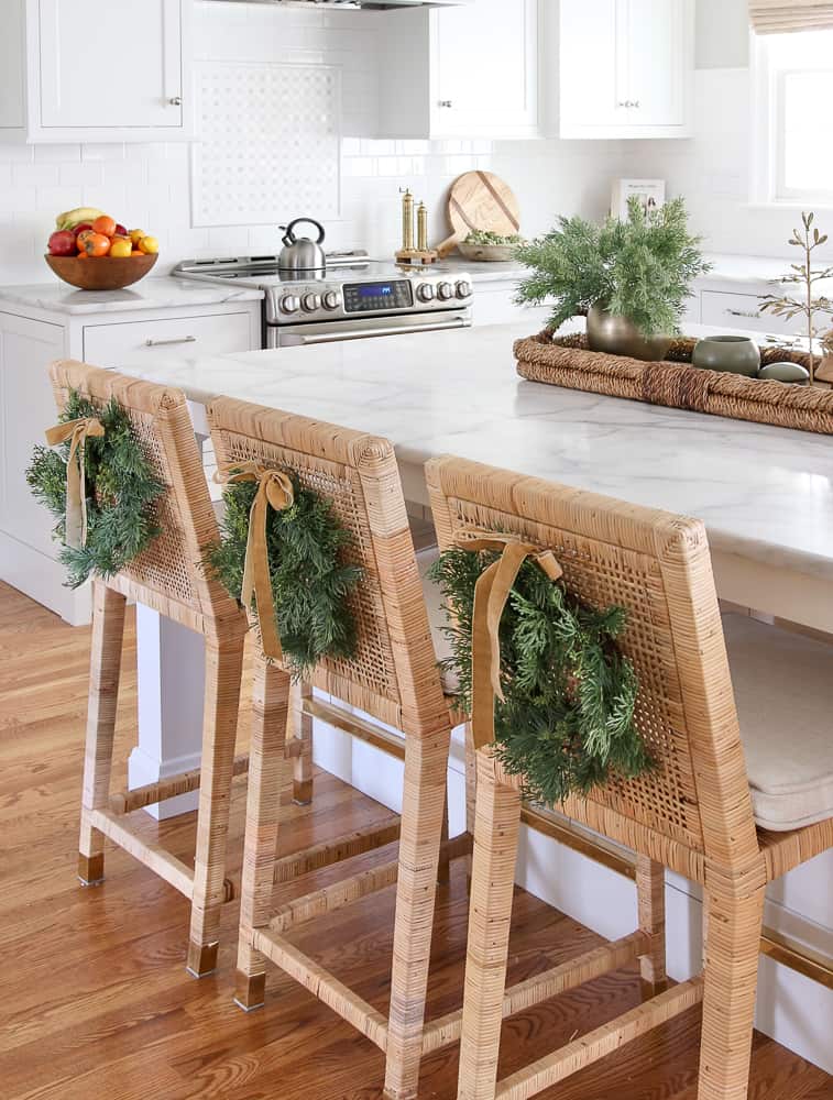 hang wreaths on the back of counter stools, white kitchen with Carrara marble countertops, rattan counter stools with small wreaths and velvet ribbon hanging on the back of the stools