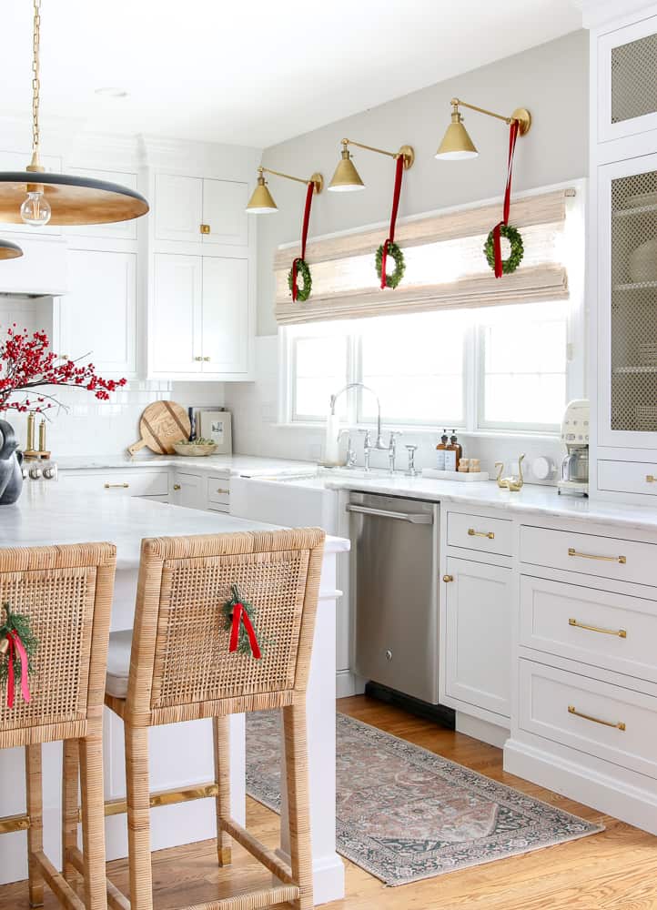 White kitchen decorated for Christmas, sconces over sink with small wreaths and ribbons, cedar sprigs on back rattan kitchen counter stools, red berry stems on island countertop