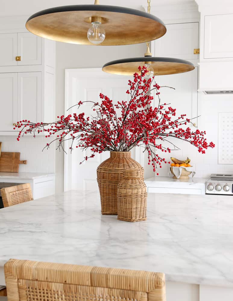 classic white kitchen decorated for Christmas, marble island countertop with wicker vases and stems with red berries, rattan counter stools