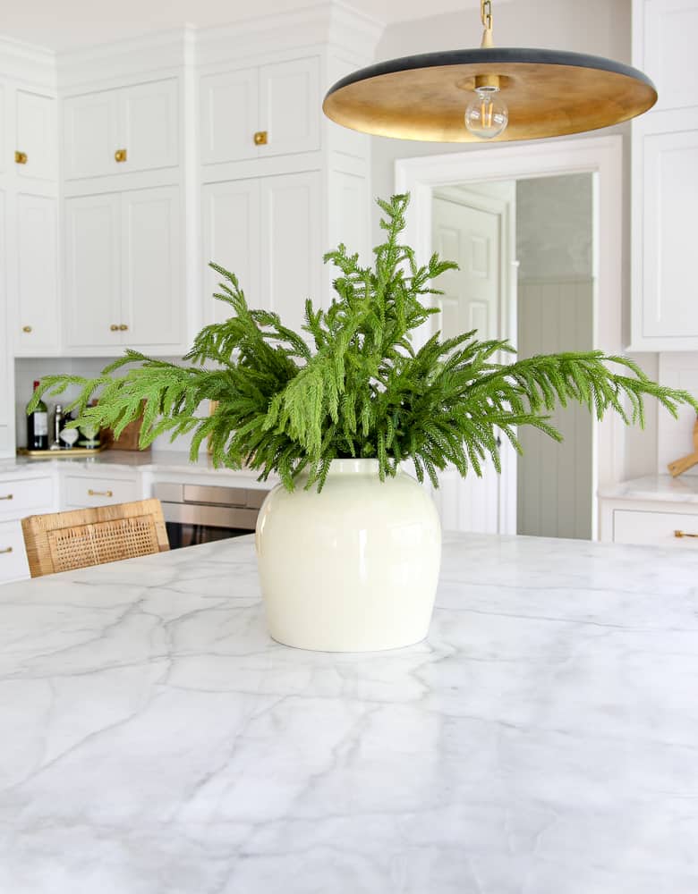 Norfolk pine branches styled in a centerpiece and a kitchen island, a classic white kitchen with unlacquered brass hardware, rattan counter stools, marble countertops