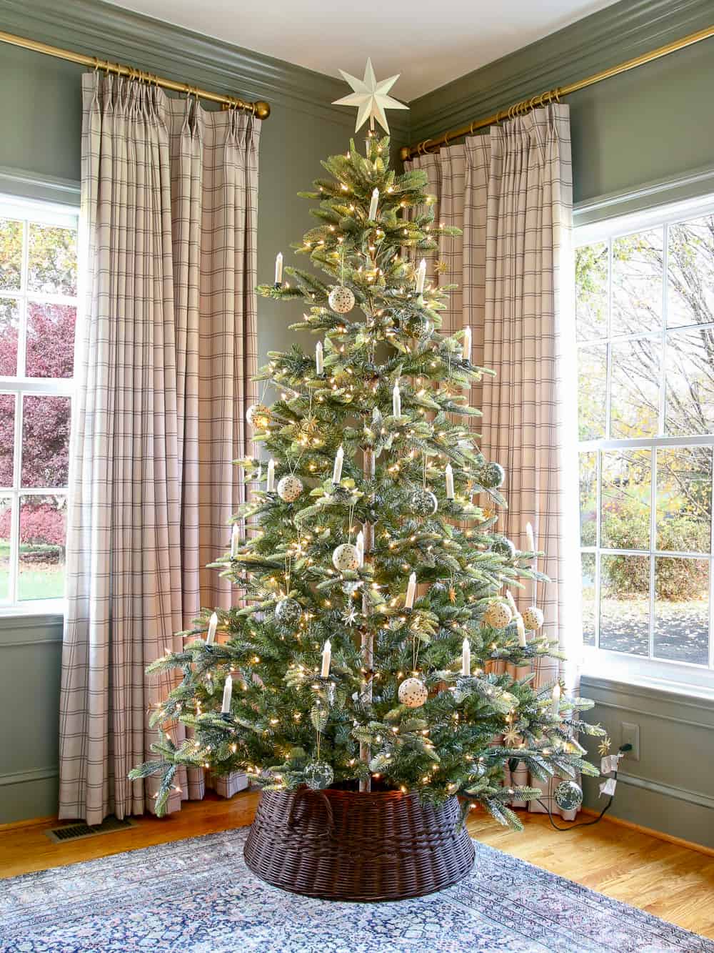 Artificial Christmas tree with vintage candles and ornaments in a dark woven tree collar, windowpane linen drapes on windows, Storm cloud gray walls, brass curtain rod, crown molding
