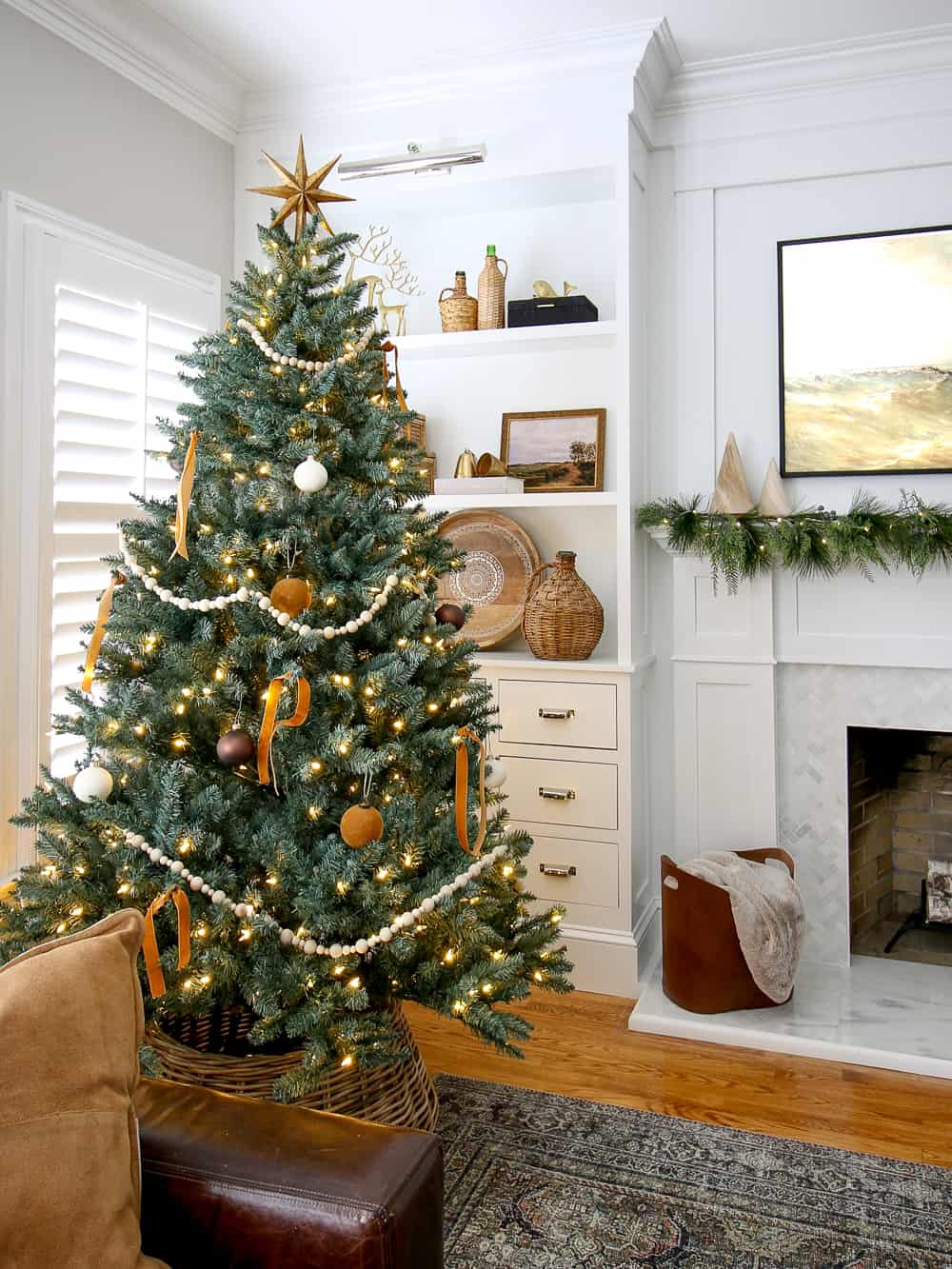 Full artificial blue spruce in living room decorated with neutral ornaments, wood bead garland and a gold star topper, frame tv over fireplace mantel decorated for Christmas with garland and wooden trees, shelf decor in background with woven elements and vintage art