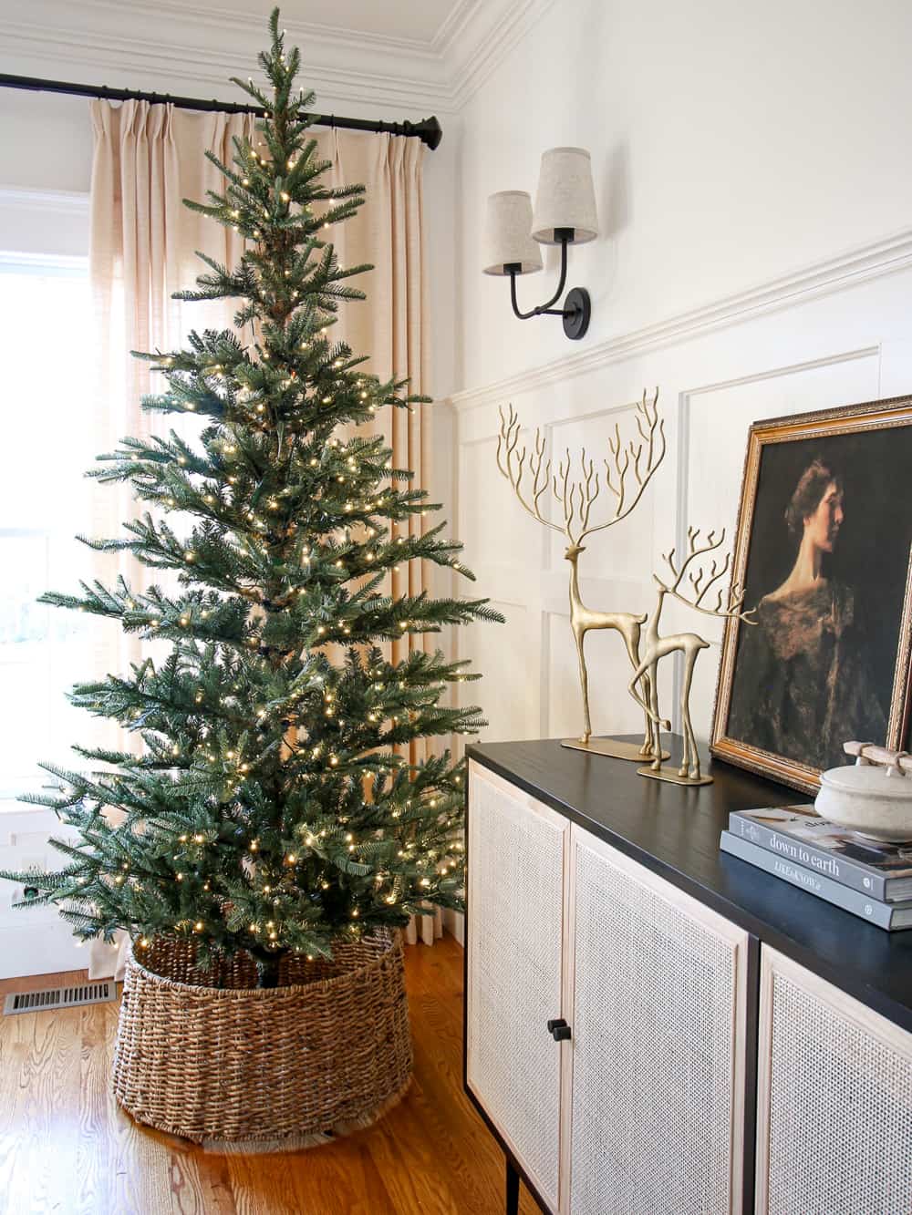 sparse Christmas tree in dining room next to cane cabinet styled with Pottery barn brass reindeer, vintage artwork, and a trinket dish top of coffee table books, wall molding, linen drapes, hardwood floors