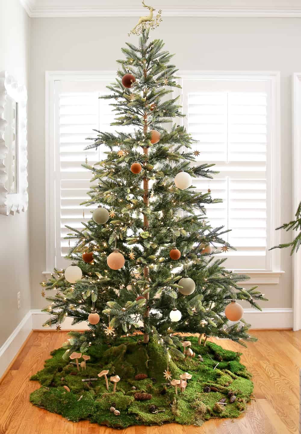 Sparse fir Christmas tree with velvet ornaments, living room with agreeable gray walls, interior window shutters, woodland theme tree skirt with moss, mushrooms, pinecones