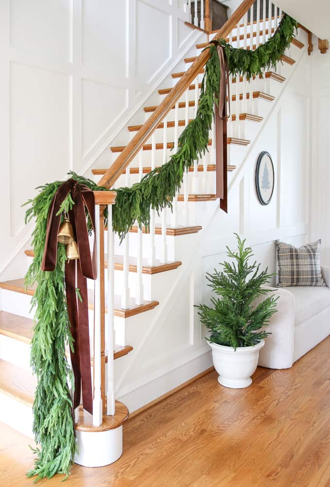 entryway hallway and staircase with garland draped down the banister, Norfolk pine garland, brown velvet ribbon, wall molding, boucle bench  styled with pillow, vintage Christmas tree wall art and small Norfolk pine tree