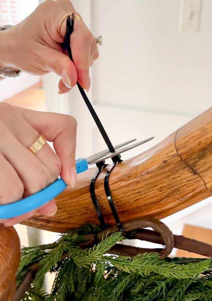 person demonstrating how use a zip tie to hang garland on a staircase and cutting excess plastic with scissors