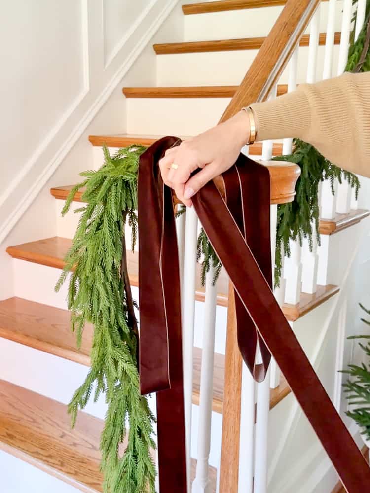 person using velvet ribbon to tie a bow as Christmas staircase decor on garland