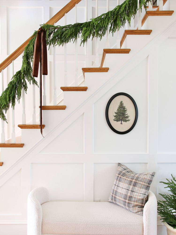white hallway with wall molding, white boucle bench, vintage Christmas tree art, throw pillow, small pine tree, garland hanging from banister