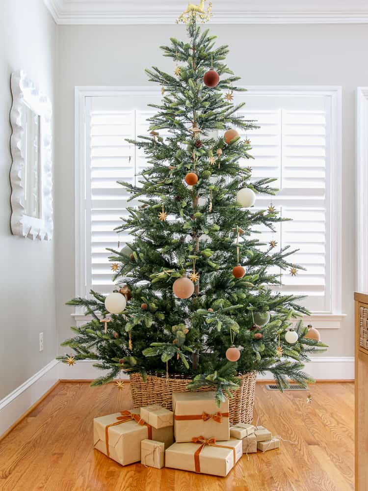 sparse tree with minimal velvet Christmas ornaments, living room with agreeable gray walls, hardwood floors, window with interior shutters, woven tree collar, gifts wrapped under the tree