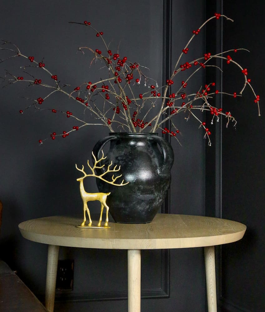 single brass reindeer styled with black vase and berry stems on round wooden end table in room with black walls