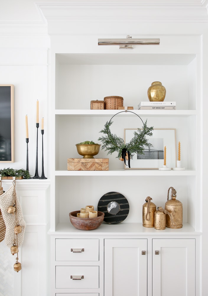 built-in shelves on fireplace wall styled for Christmas, vintage brass bells, simple wreath, candles, storage box, round woven boxes brass objects, picture light
