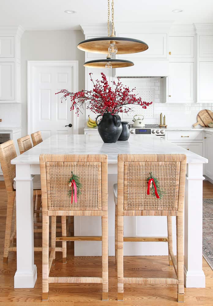 Minimalist Christmas kitchen decor, sprigs and ribbon on the back of rattan counter stools, red berry stems  styled in a black vase on honed marble countertops in white kitchen with brass hardware 