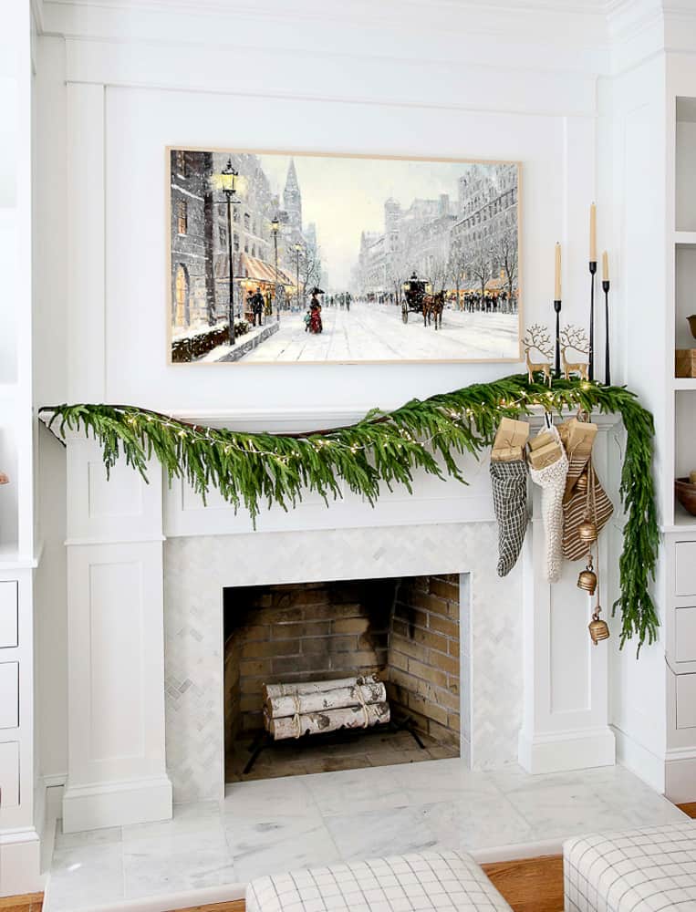 fireplace mantel decorated for christmas, Norfolk pine garland, fairy lights, stockings and bells, frame tv with winter art, tapered candle holders, birch logs in fireplace