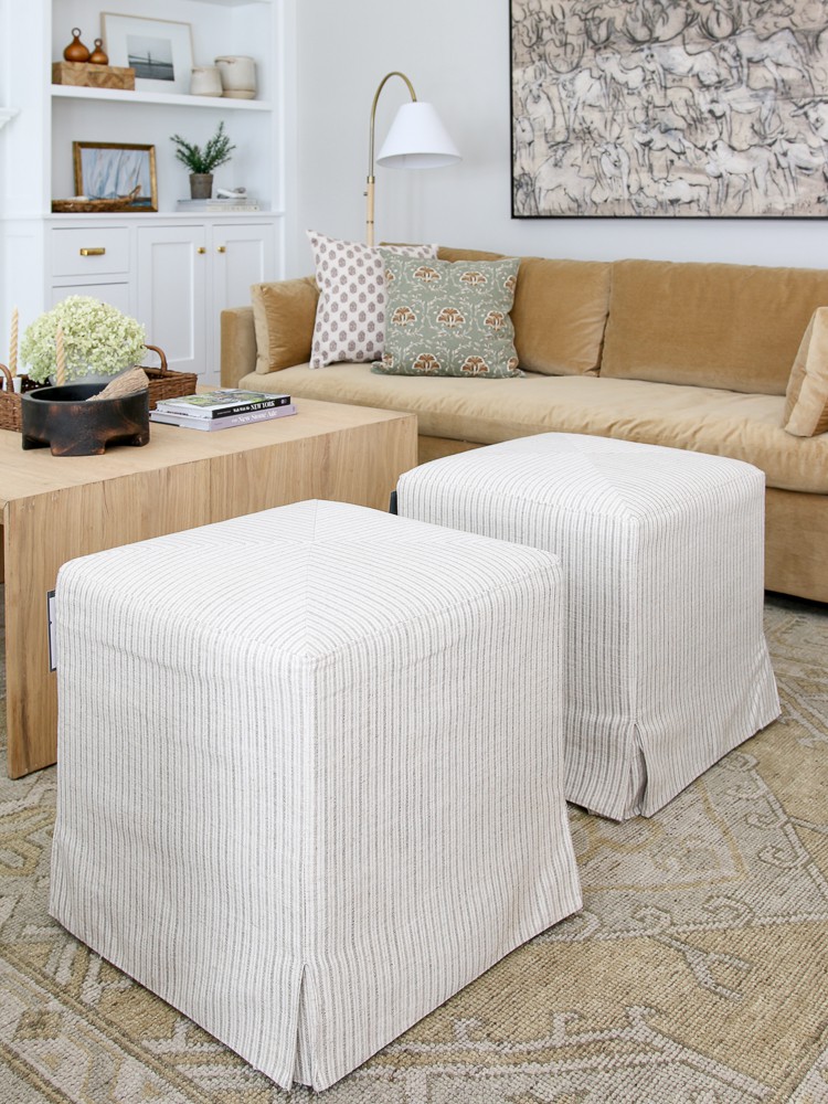 two ottomans styled in a living room with a hand knotted rug, velvet camel colored sofa, square wood coffee table 