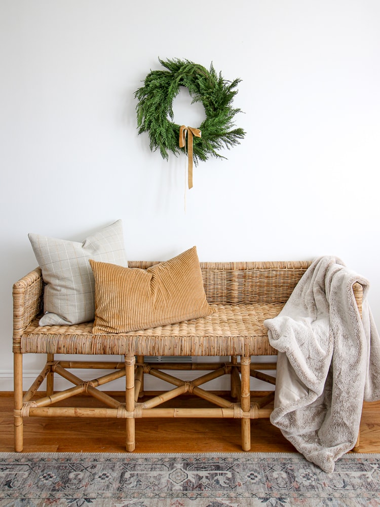 hang wreaths on a wall above furniture, Christmas cypress wreath hanging on a white wall above a woven bench, camel colored ribbon coordinates with pillows and throw blanket styled on bench