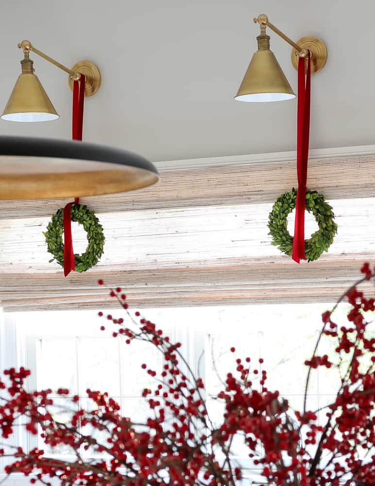 hang wreaths from light fixtures, close up of small boxwood wreaths hung with a red velvet ribbon from brass sconces above a window in a kitchen
