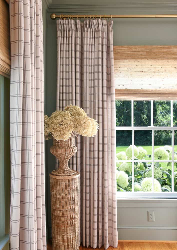 corner of home office with greenish gray walls, Roman woven shades, windowpane drapes, wicker pedestal and urn with dried hydrangeas