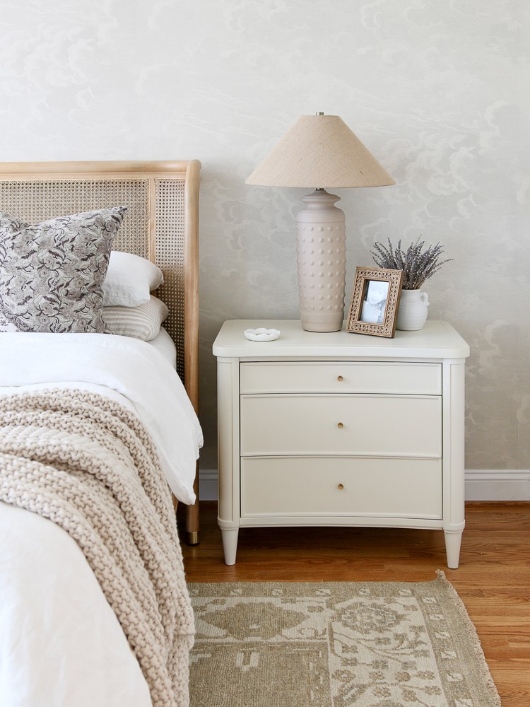 nightstand styling with a lamp, photo frame and plant, neutral bedroom with cane bed, white nightstand and nuvolette wallpaper, hand knotted rug, nightstand decor